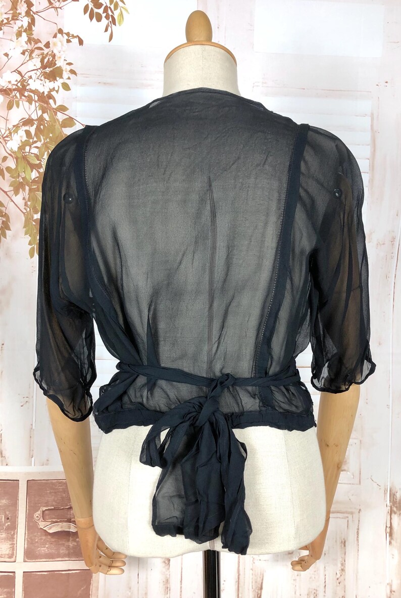 May include: Black sheer bolero top with short sleeves and a tie closure at the waist. The bolero is displayed on a mannequin, showcasing its delicate, flowing fabric and design. The top has a ruffled hem and is made of a lightweight material.