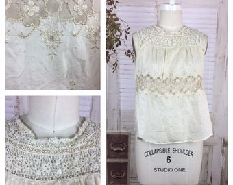 May include: A white sleeveless blouse with delicate lace detailing around the neckline and hem. The blouse has a gathered, slightly puffed look and is made of a lightweight fabric.