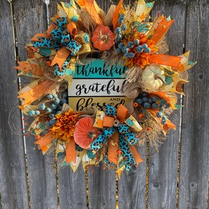May include: A fall-themed wreath with orange, yellow, and blue accents. It features a sign that reads "thankful grateful and blessed".
