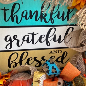 May include: A wooden sign with the words "thankful grateful and blessed" in black script on a turquoise, white, and gold background. The sign is decorated with burlap, orange, blue, and leopard print ribbon, and a small pumpkin.