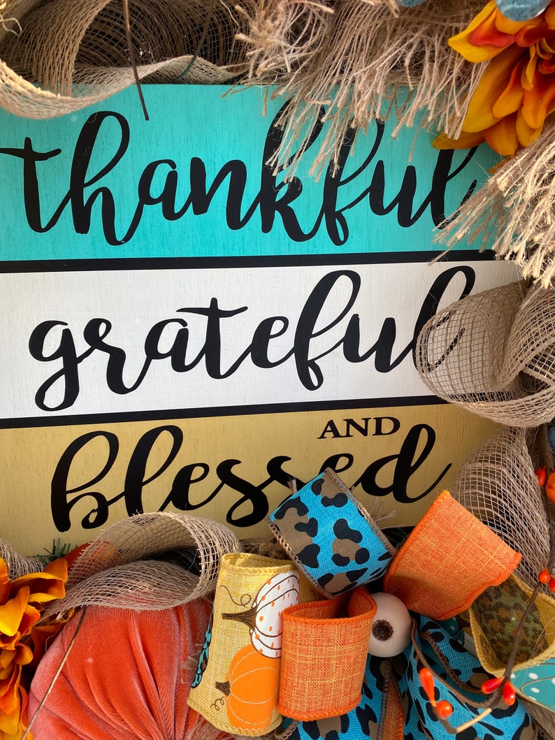 May include: A wooden sign with the words "thankful grateful and blessed" in black script on a turquoise, white, and gold background. The sign is decorated with burlap, orange, blue, and leopard print ribbon, and a small pumpkin.