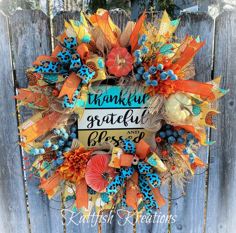 May include: A fall-themed wreath with orange, blue and brown accents. It features a sign that reads "thankful grateful and blessed".