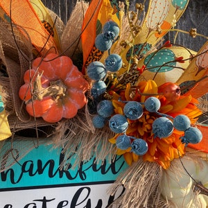 May include: A fall-themed wreath with a turquoise and yellow sign that reads "thankful grateful and blessed". The wreath is decorated with orange and white pumpkins, burlap, blue berries, and a yellow and orange floral arrangement.