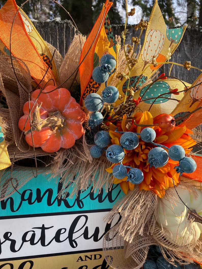 May include: A fall-themed wreath with a turquoise and yellow sign that reads "thankful grateful and blessed". The wreath is decorated with orange and white pumpkins, burlap, blue berries, and a yellow and orange floral arrangement.