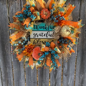 May include: A fall-themed wreath with orange, yellow, and blue ribbons, faux pumpkins, and a sign that reads "thankful grateful and blessed".