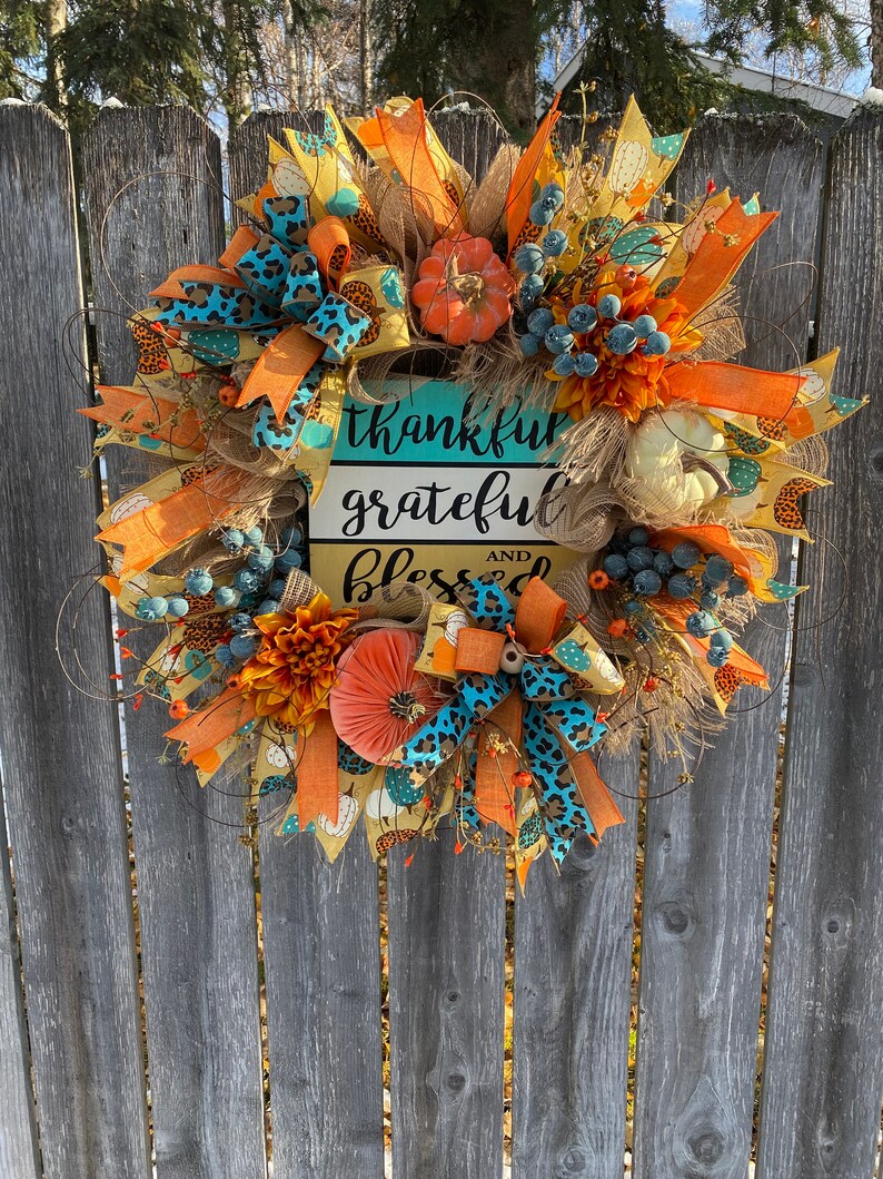 May include: A fall-themed wreath with orange, yellow, and blue ribbons, faux pumpkins, and a sign that reads "thankful grateful and blessed".