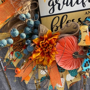 May include: A fall-themed wreath with a sign that reads "grateful and blessed". The wreath is decorated with orange, brown, and blue ribbons, a velvet pumpkin, and a large orange flower.