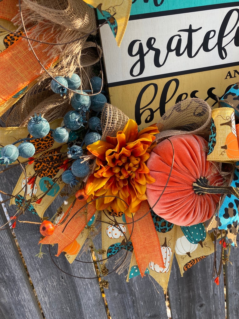 May include: A fall-themed wreath with a sign that reads "grateful and blessed". The wreath is decorated with orange, brown, and blue ribbons, a velvet pumpkin, and a large orange flower.