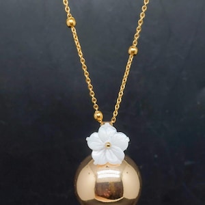 May include: A gold-colored necklace with a round pendant and a white flower accent. The necklace has a delicate chain with small gold beads. The pendant is a shiny sphere. The flower is white with a small gold center.