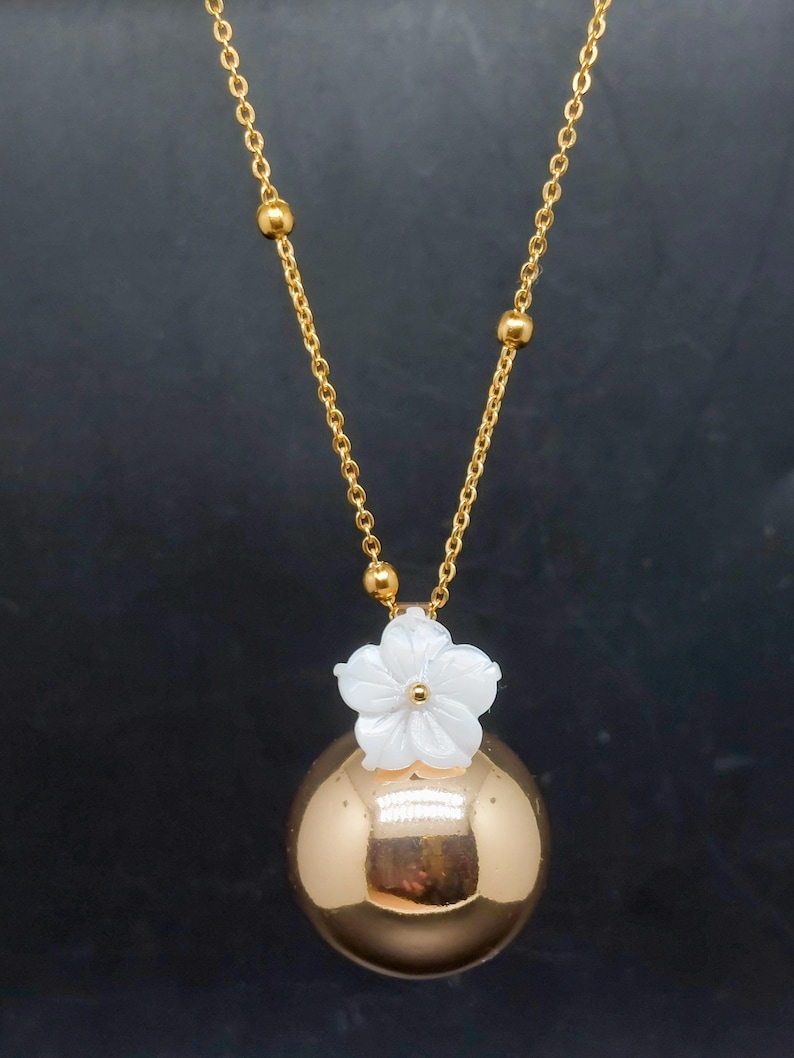 May include: A gold-colored necklace with a round pendant and a white flower accent. The necklace has a delicate chain with small gold beads. The pendant is a shiny sphere. The flower is white with a small gold center.