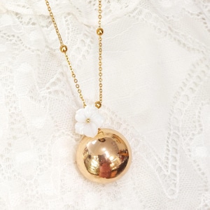 May include: A gold-colored necklace with a large, round pendant. The pendant is accented with a small white flower. The necklace chain is delicate, with small gold beads interspersed along its length. The background is a white, lacy fabric.