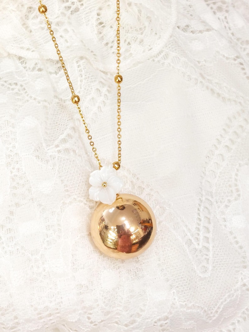 May include: A gold-colored necklace with a large, round pendant. The pendant is accented with a small white flower. The necklace chain is delicate, with small gold beads interspersed along its length. The background is a white, lacy fabric.