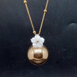 May include: A gold-colored necklace featuring a large, spherical pendant. A white flower sits atop the sphere. The necklace chain is gold with small, round accents. The background is a dark, solid color.