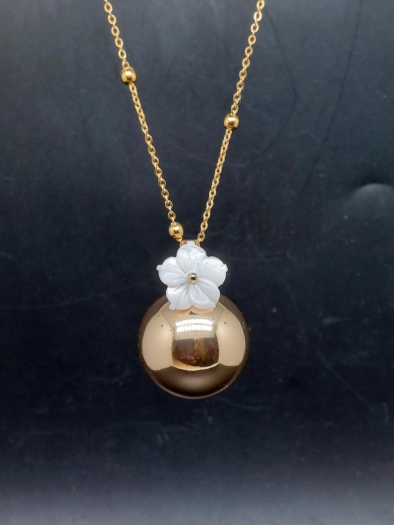 May include: A gold-colored necklace featuring a large, spherical pendant. A white flower sits atop the sphere. The necklace chain is gold with small, round accents. The background is a dark, solid color.