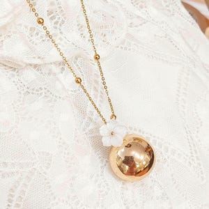 Peut inclure: Un collier doré avec un grand pendentif rond. Le pendentif est orné d'une petite fleur blanche. La chaîne est fine, avec de petites perles dorées. Le fond est un tissu blanc et dentelle, créant un contraste délicat.