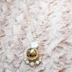 Pu&ograve; includere: Una collana d'oro con un grande ciondolo a forma di sfera dorata circondato da un cerchio di perle bianche e un ciondolo a forma di fiore bianco.