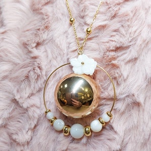 Pu&ograve; includere: Una collana d'oro con un grande ciondolo rotondo in oro e un ciondolo a forma di fiore bianco. Il ciondolo &egrave; circondato da un cerchio di perle bianche.