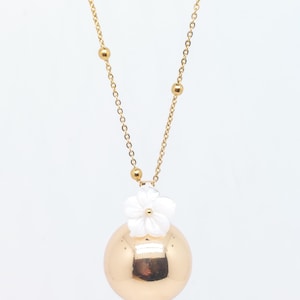 May include: A gold-colored necklace with a large, round pendant. A white flower is attached to the top of the pendant. The necklace chain is gold and features small gold beads.