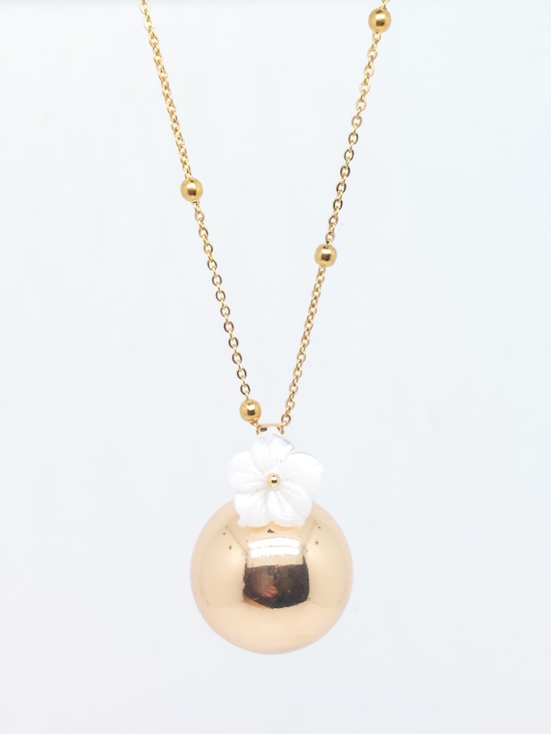 May include: A gold-colored necklace with a large, round pendant. A white flower is attached to the top of the pendant. The necklace chain is gold and features small gold beads.