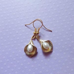 May include: A pair of gold-colored earrings featuring a calla lily design. Each earring has a gold-colored lily with a white pearl center, suspended from a gold-colored hook. The earrings are displayed against a light purple background.
