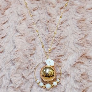 Pu&ograve; includere: Una collana d'oro con un grande ciondolo sferico dorato circondato da un cerchio di perline bianche e un piccolo ciondolo a forma di fiore bianco.