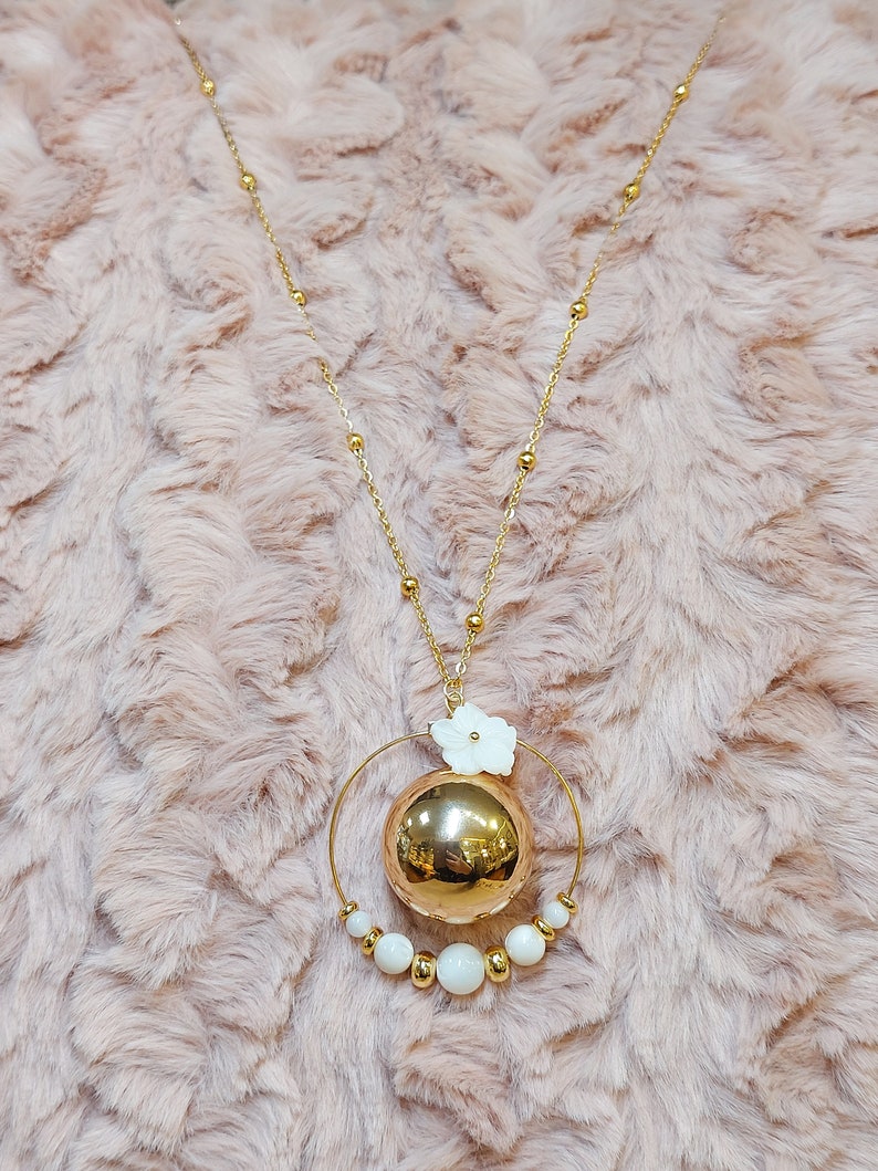 Pu&ograve; includere: Una collana d'oro con un grande ciondolo sferico dorato circondato da un cerchio di perline bianche e un piccolo ciondolo a forma di fiore bianco.