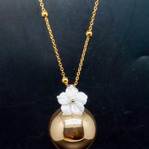 May include: A gold-colored necklace featuring a spherical pendant with a white flower accent. The pendant hangs from a delicate gold chain with small, round beads. The necklace is displayed against a dark background.