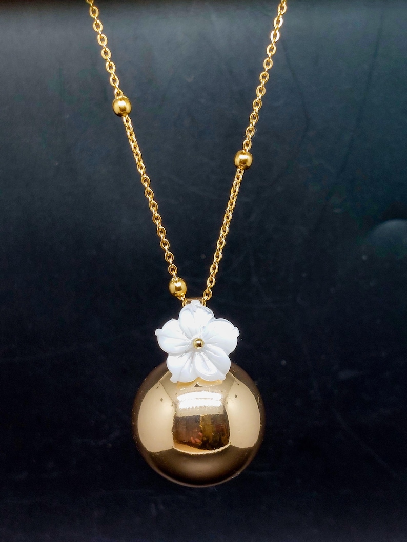 May include: A gold-colored necklace featuring a spherical pendant with a white flower accent. The pendant hangs from a delicate gold chain with small, round beads. The necklace is displayed against a dark background.