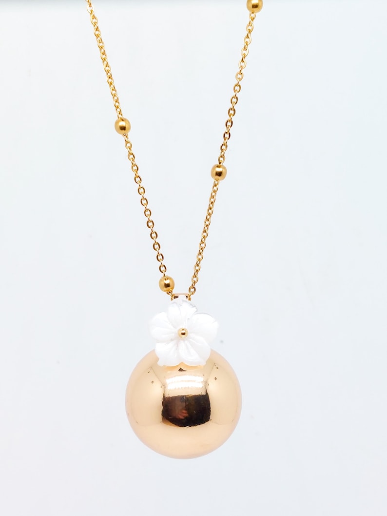 May include: Gold-toned necklace featuring a large, round pendant with a white flower accent. The chain has small gold beads and a delicate link design. The pendant reflects light, creating a polished look. A stylish piece of jewelry.
