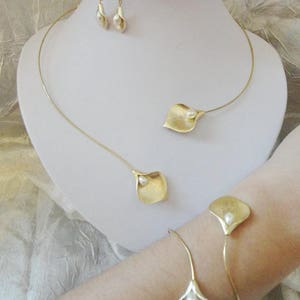 May include: A gold-toned jewelry set featuring a necklace, earrings, and bracelet. The necklace and bracelet have a delicate, flowing design with a calla lily motif. Each piece includes a small, white pearl accent. The earrings are also calla lily shaped.