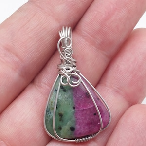 Puede incluir: Un colgante de piedra preciosa verde y rosa envuelto en alambre de plata. El colgante tiene forma de lágrima y tiene un aro de plata.