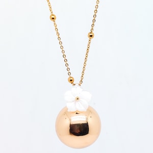 May include: A gold-colored necklace featuring a large, spherical pendant. A white flower adorns the top of the pendant. The necklace chain is gold and includes small, round beads. The jewelry is set against a white background.