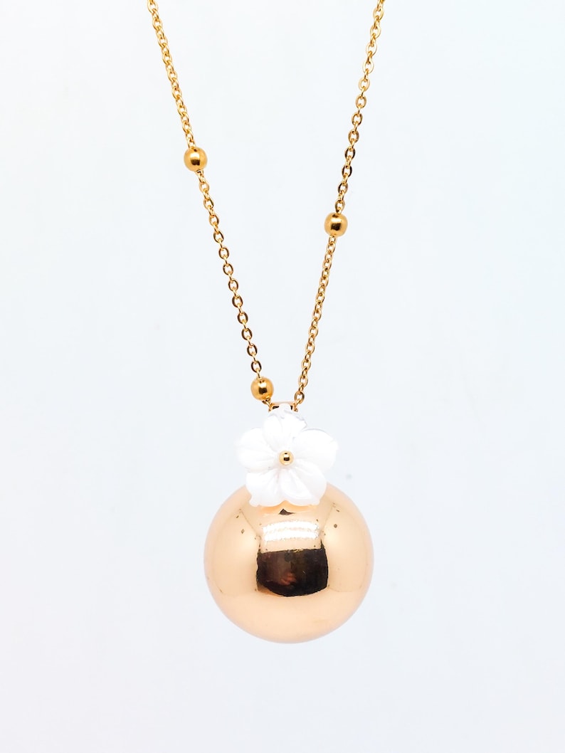 May include: A gold-colored necklace featuring a large, spherical pendant. A white flower adorns the top of the pendant. The necklace chain is gold and includes small, round beads. The jewelry is set against a white background.