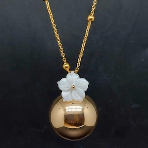 May include: A gold-colored necklace featuring a round pendant with a white flower accent. The chain is also gold, with small gold beads. The pendant is a reflective, spherical shape. The necklace is displayed against a dark background.
