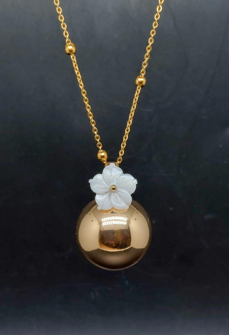 May include: A gold-colored necklace featuring a round pendant with a white flower accent. The chain is also gold, with small gold beads. The pendant is a reflective, spherical shape. The necklace is displayed against a dark background.