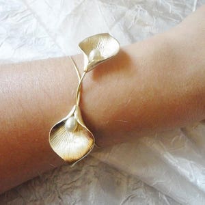 May include: A gold-toned bracelet featuring two calla lily flower designs, each with a pearl center. The bracelet is worn on a wrist, showcasing the intricate details of the floral design. The bracelet is made of metal.