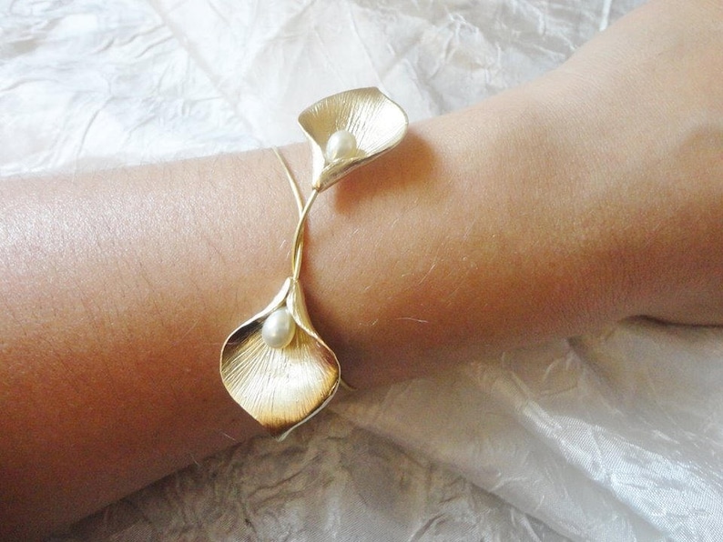 May include: A gold-toned bracelet featuring two calla lily flower designs, each with a pearl center. The bracelet is worn on a wrist, showcasing the intricate details of the floral design. The bracelet is made of metal.