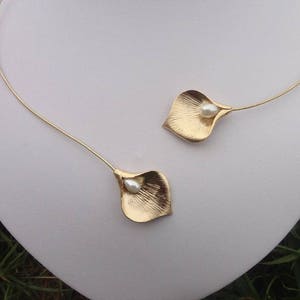 May include: A gold-colored necklace featuring two stylized calla lily pendants. Each pendant has a textured surface and a small, white pearl at its center. The necklace is displayed on a white mannequin.