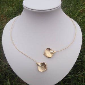 May include: A gold-colored necklace with a delicate design. The necklace features a thin, curved wire that forms a choker. Two gold-colored, petal-shaped pendants, each with a small white pearl, are attached to the wire.