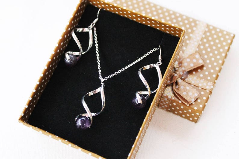 Op de afbeelding: Een zilveren ketting en oorbellen set met een gedraaid ontwerp en een hanger met een paarse edelsteen. De sieraden worden gepresenteerd in een bruine doos met witte stippen.