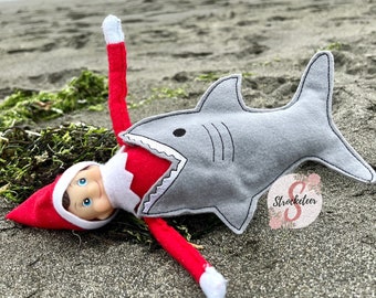 May include: A whimsical felt shark toy with a red and white elf doll partially inside, lying on a sandy beach. The shark is gray with black stitched details. The elf is wearing a red hat and suit. The scene is set on a beach with seaweed.