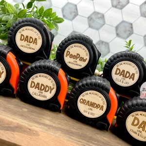 May include: A collection of personalized black and orange tape measures with wooden engraved labels. The labels read "DADA," "Daddy," "Grandpa," and "Dad," each with a name or names below. The text "No one measures up to you" is also present.