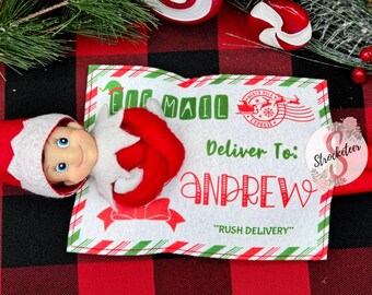 May include: A Christmas elf doll with a red and white hat and outfit, lying on a white felt letter. The letter reads "AIR MAIL" and "Deliver To: ANDREW" with a North Pole Express stamp. The background is a red and black plaid pattern.