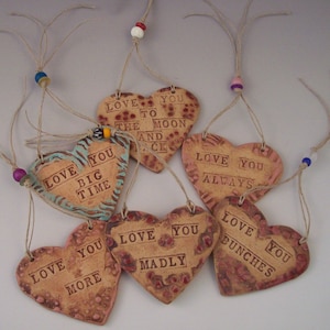 May include: Eight handmade ceramic heart ornaments with different love-themed messages, each hanging from a twine string with a colourful bead. The messages include "Love You To The Moon And Back", "Love You Big Time", "Love You More", "Love You Madly", "Love You Always", "Love You Bunches".