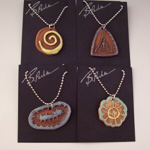 May include: Four ceramic pendants on silver chains, each with a different design: a spiral, a triangle, a flower, and a wavy line. The pendants are displayed on black cards with the signature "B. Parker" on each.