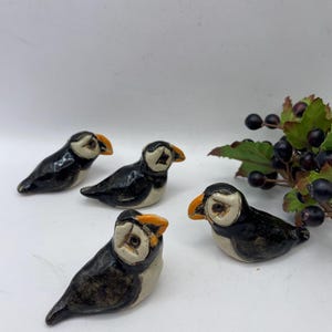 May include: Four ceramic puffin figurines. Each bird has a black body, white face, and an orange beak. The puffins are arranged on a white surface, with a sprig of berries in the background.