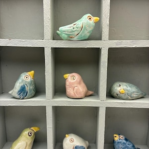 Könnte beinhalten: Sieben kleine Keramikvogel-Figuren in verschiedenen Farben wie Minzgrün, Hellblau, Rosa, Blassgelb, Weiß und Dunkelblau. Jeder Vogel hat eine glänzende Oberfläche, einen kleinen gelben Schnabel und aufgemalte Augen, platziert in einzelnen grauen Fächern.