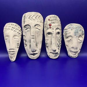 May include: Four white, carved face masks with closed eyes, arranged on a blue background. Each mask has unique details, including textured patterns and subtle color accents. The masks vary in size and shape, showcasing a handcrafted aesthetic.