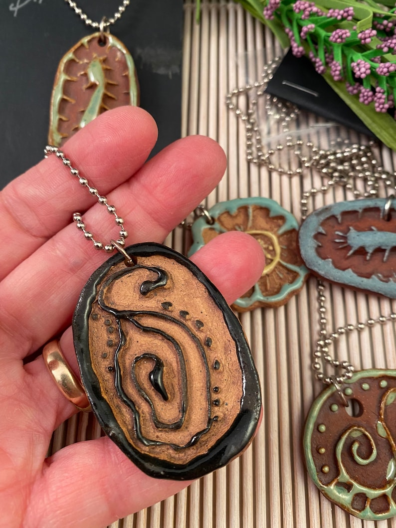 May include: A set of four ceramic pendants with different designs. Each pendant is brown with a black outline and features a unique pattern. The pendants are on silver chains.