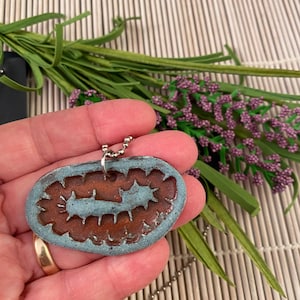 May include: A blue and brown ceramic pendant with a detailed design, hanging from a silver chain. The pendant is shaped like a worm or caterpillar.